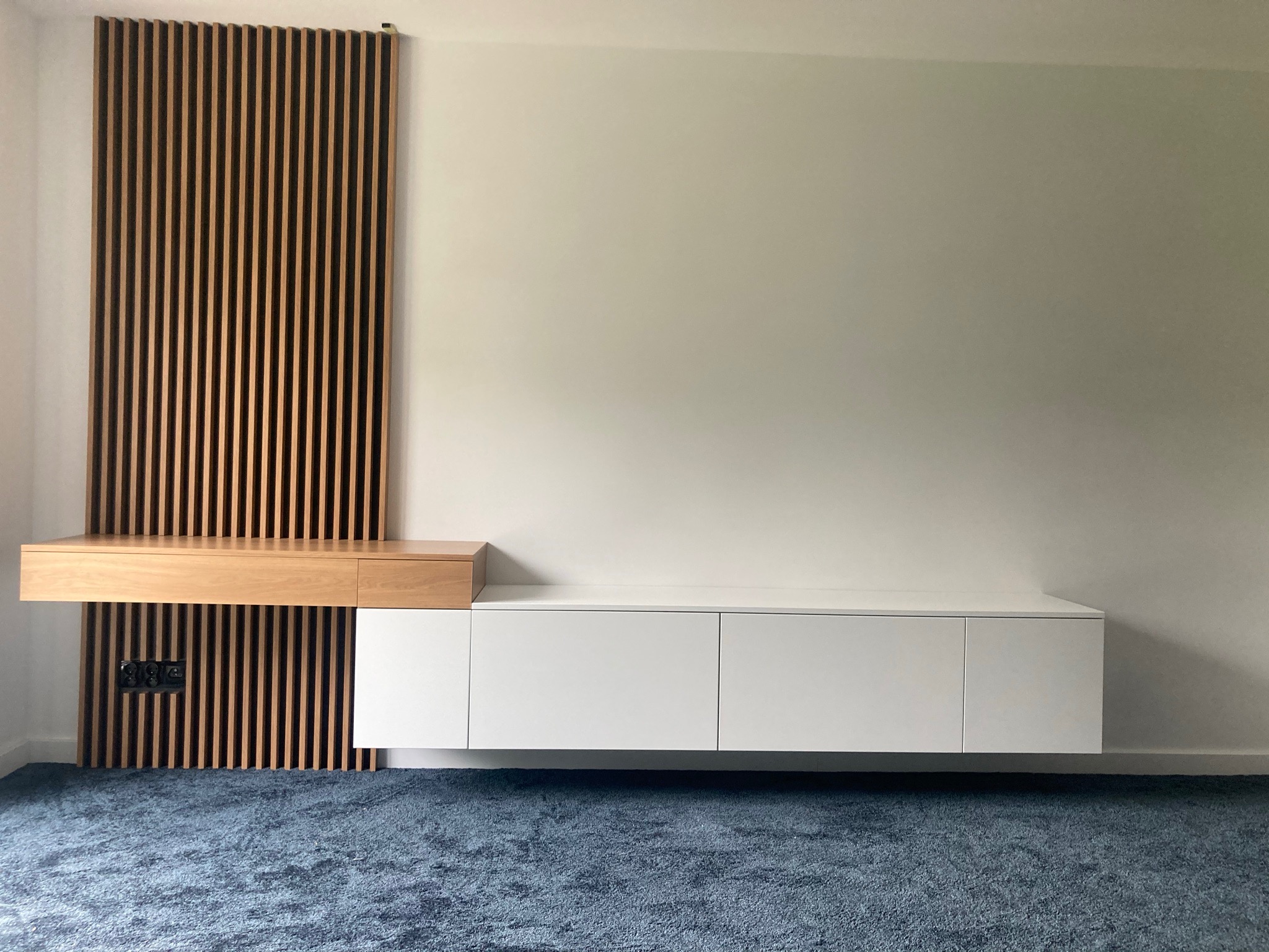 Nowoczesna zabudowa ściany z drewnianymi lamelami, wiszącą szafką RTV z białymi frontami i drewnianą półką z szufladą, na tle błękitnej wykładziny dywanowej.