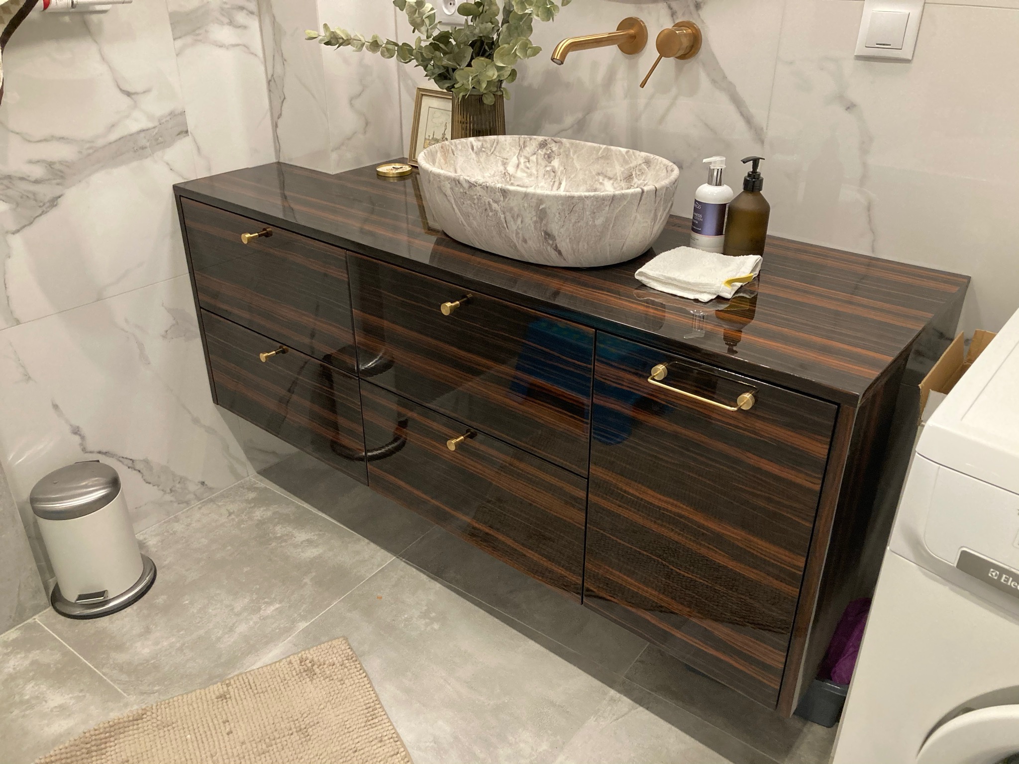 Łazienka z blatem z ciemnego drewna egzotycznego, na którym umieszczona jest marmurowa umywalka w kształcie misy, złota bateria ścienna i akcesoria łazienkowe.