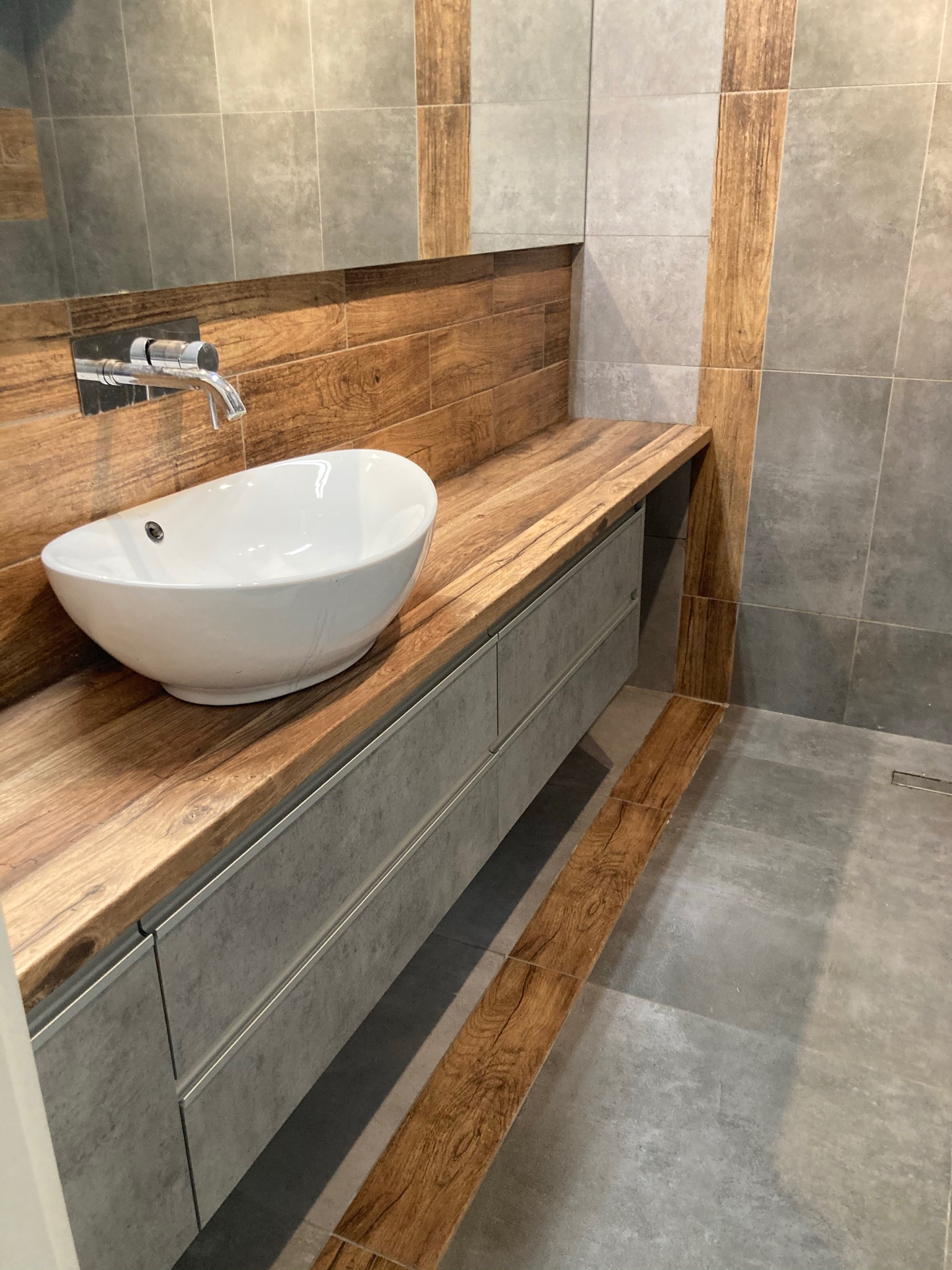 Nowoczesna łazienka z umywalką nablatową na drewnianym blacie i szarą szafką z szufladami, płytki w odcieniach szarości z drewnianymi akcentami.