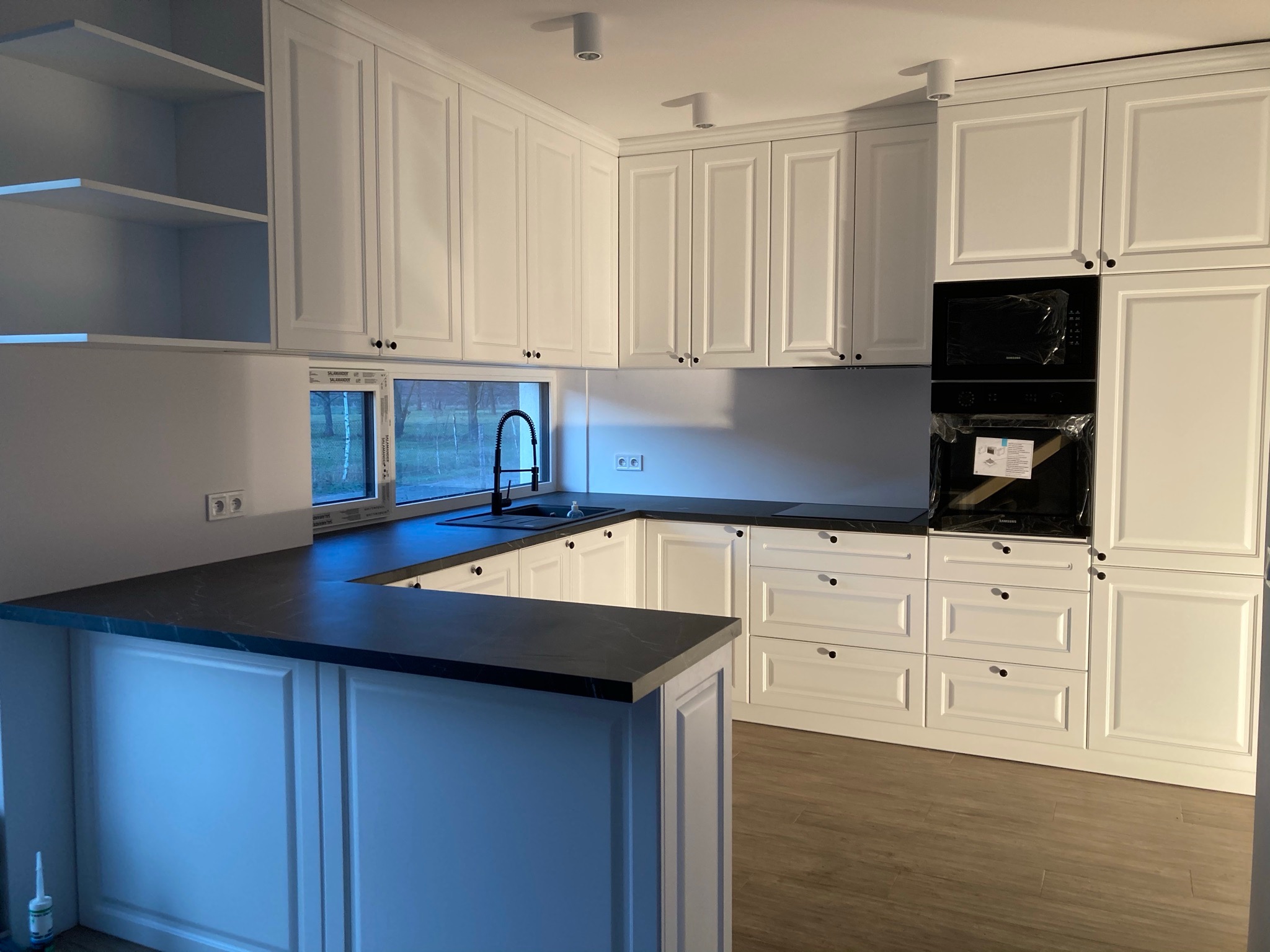 Nowoczesna kuchnia w bieli z ciemnym blatem, widok na okno i zabudowany piekarnik w folii ochronnej.