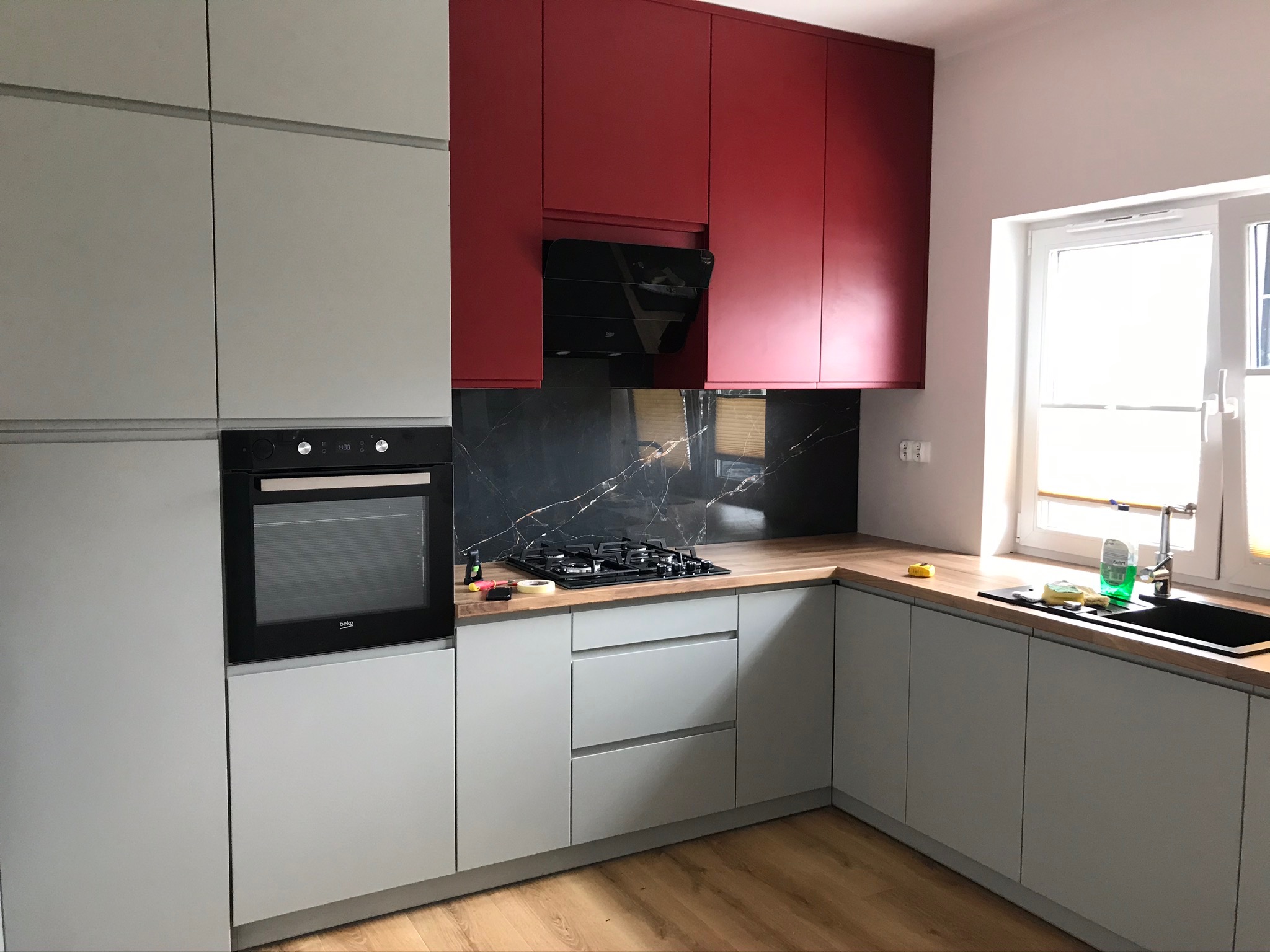 Szare szafki kuchenne z czerwonymi szafkami wiszącymi i czarnym blatem z marmurowym wzorem, widoczny piekarnik i płyta gazowa.