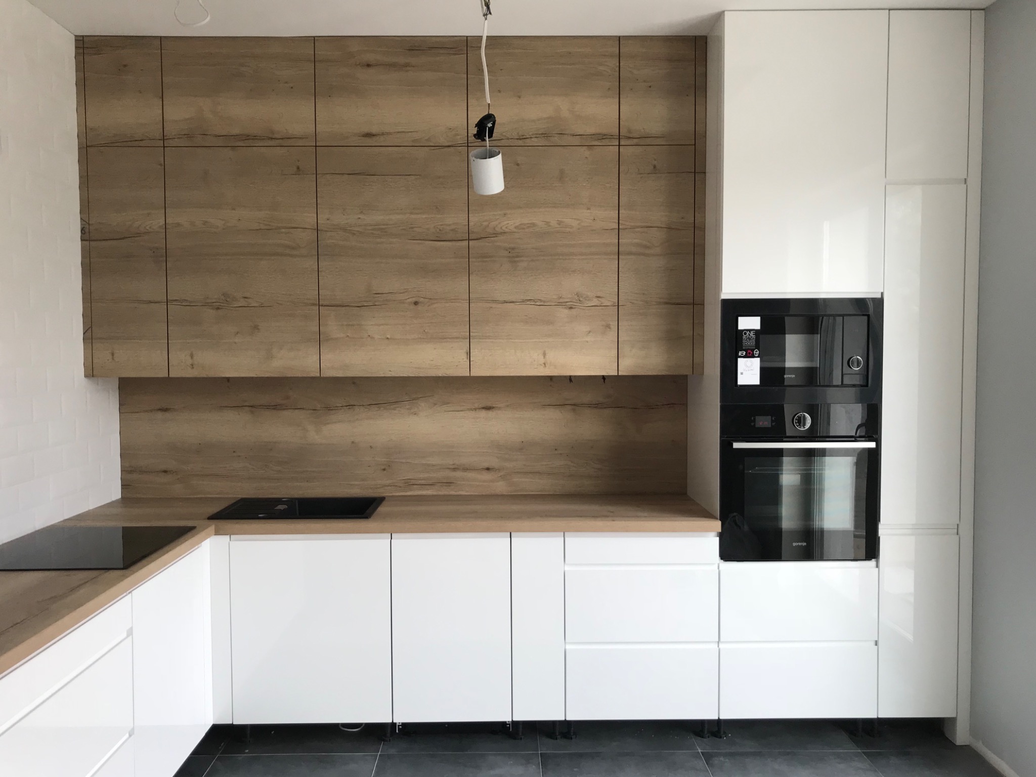 Nowoczesna kuchnia z białymi szafkami na wysoki połysk i drewnianymi panelami górnymi, czarna płyta indukcyjna i piekarnik w zabudowie, wisząca lampa bez klosza.