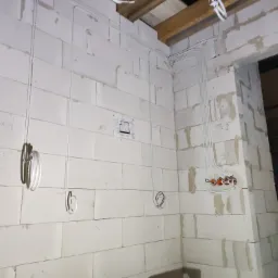 Wewnętrzna instalacja elektryczna w surowym stanie, białe przewody poprowadzone po ścianie z bloczków, widoczne puszki elektryczne i zwisające pętle kabli.