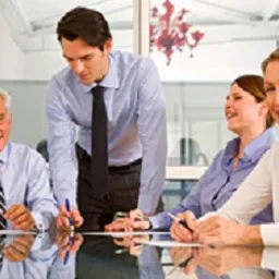 Spotkanie zespołu przy szklanym stole; mężczyzna w koszuli i krawacie pochyla się nad dokumentami, pozostali uczestnicy uśmiechnięci, w tle biurowe wnętrze z dekoracyjnym żyrandolem.