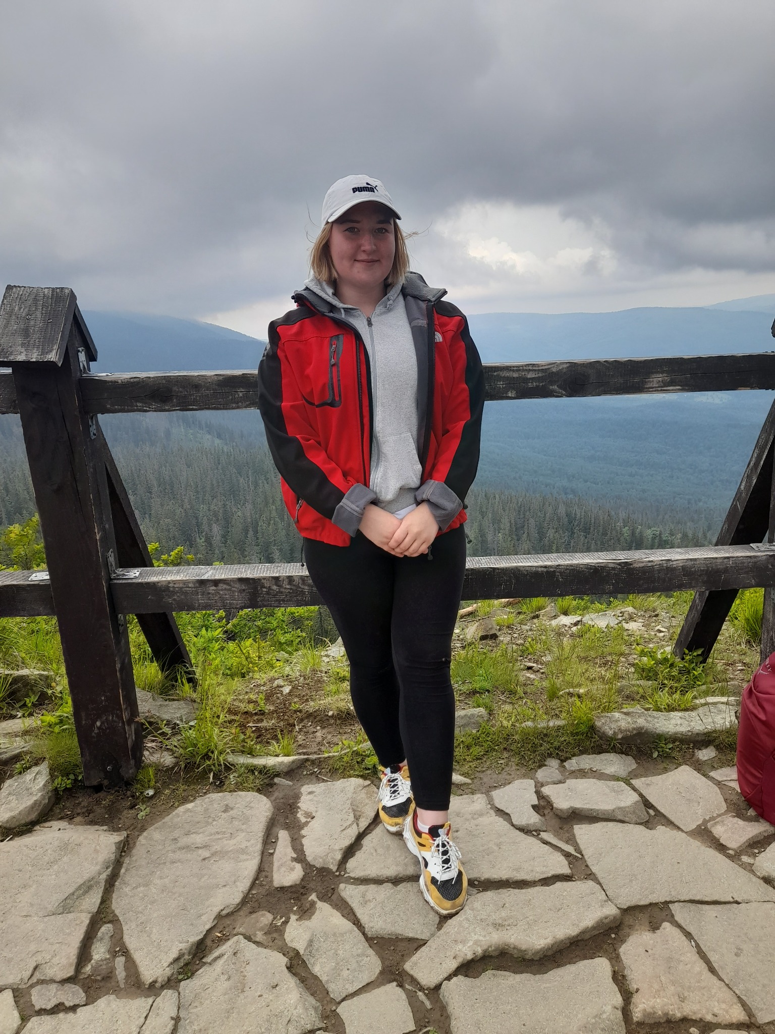 Kobieta w czerwonej kurtce sportowej i czarnych legginsach stoi na kamiennej ścieżce przy drewnianej barierce z widokiem na zalesione wzgórza w pochmurny dzień.