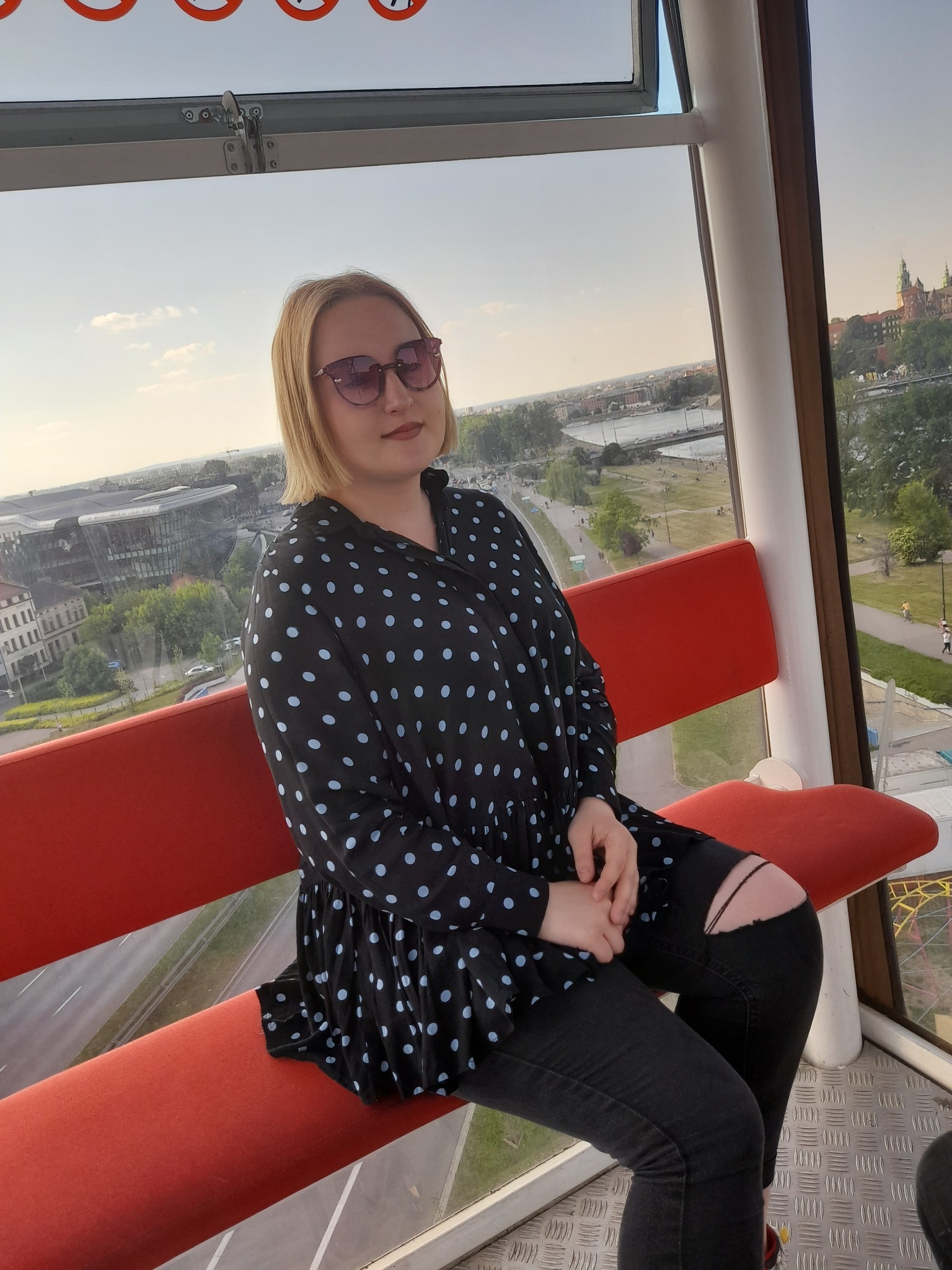 Kobieta w bluzce w grochy i podartych dżinsach siedzi na czerwonej ławce w kabinie diabelskiego młyna z widokiem na panoramę Krakowa.