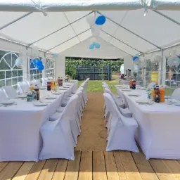 Uroczysty biały namiot z drewnianą podłogą, stoły z białymi obrusami i krzesłami w pokrowcach, dekoracje z białych i niebieskich balonów, napoje w szklanych butelkach i kwiaty w wazonach na stołach.