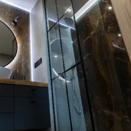Nowoczesna łazienka z kabiną prysznicową typu walk-in, marmurowymi płytkami na ścianach, oświetleniem LED i umywalką nablatową na ciemnej szafce.