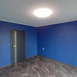 Stachu ReMake - Pomalowane na niebiesko wnętrze pokoju z szarą podłogą imitującą marmur, biały sufit z okrągłą lampą, szare drzwi z złotymi paskami i złotą klamką.