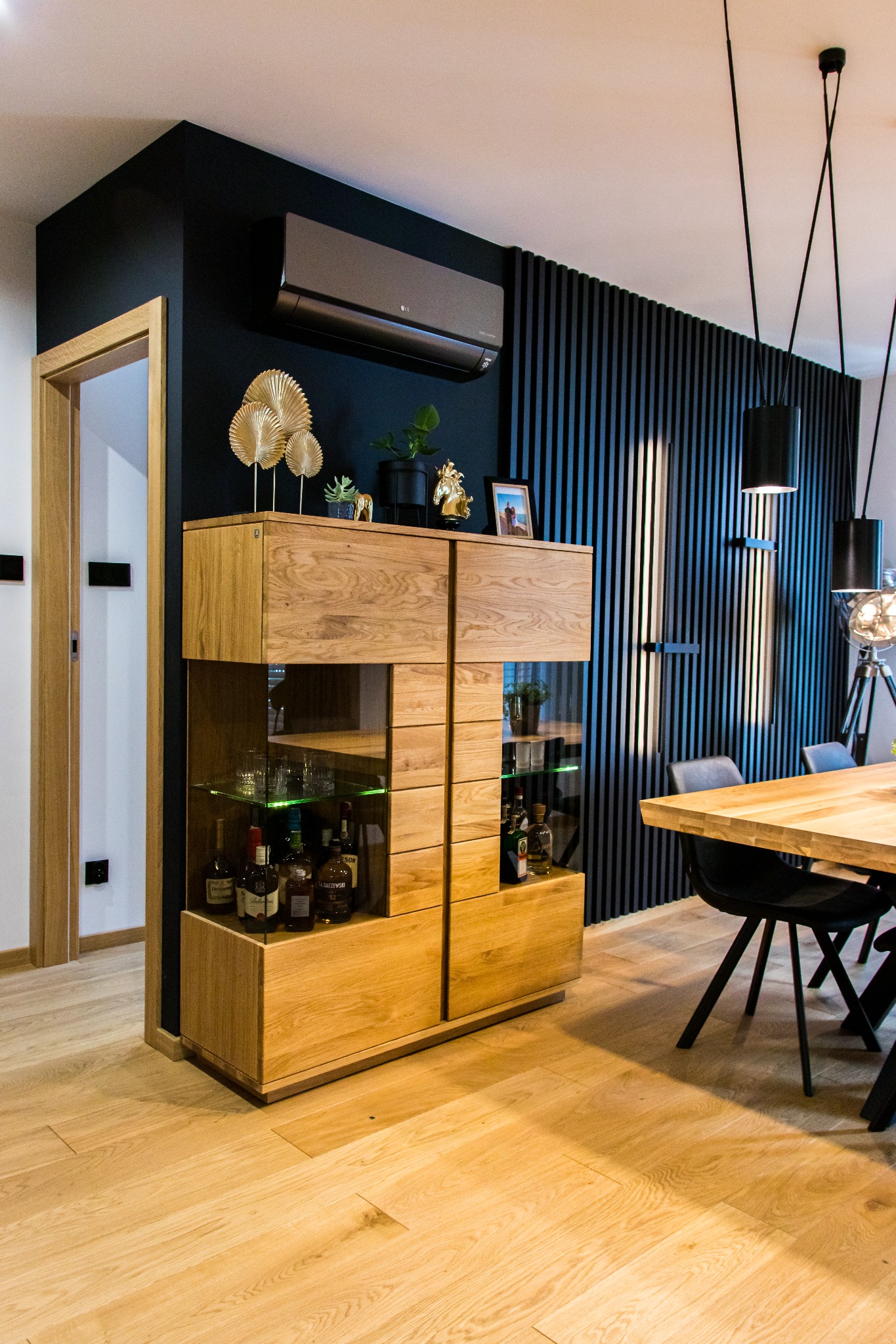 Nowoczesne wnętrze jadalni z drewnianą podłogą, szafką z przeszkleniami, czarną ścianą z lamelami i klimatyzatorem LG na górze ściany.