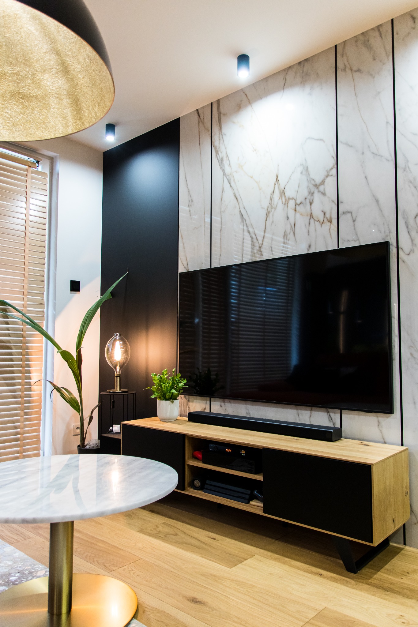 Nowoczesny salon z marmurową ścianą, telewizorem na szafce RTV z drewnianymi elementami i czarnymi drzwiczkami, okrągłym stolikiem z marmurowym blatem i złotą podstawą oraz lampą z żarówką Edisona.