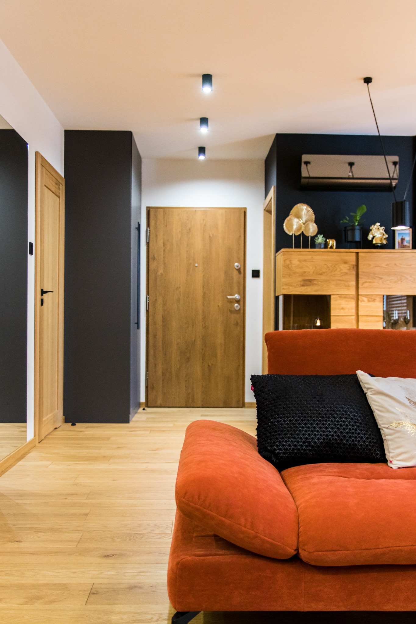 Nowoczesny salon z pomarańczową sofą, drewnianą podłogą i elementami dekoracyjnymi w odcieniach czerni i złota, oświetlony punktowymi lampami sufitowymi.