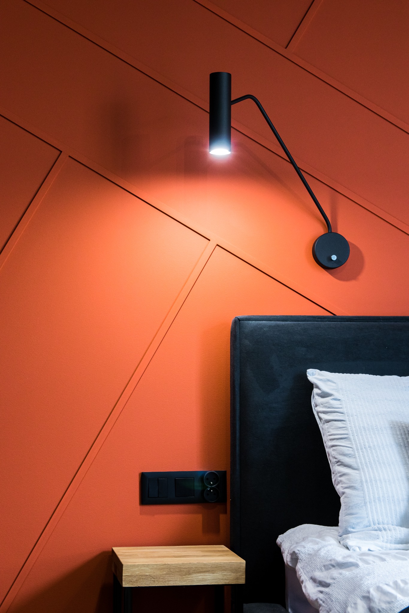 Pomarańczowa ściana z geometrycznymi panelami, oświetlona czarną lampką nocną nad wezgłowiem łóżka z czarnej tkaniny i białą pościelą, obok czarny włącznik światła.