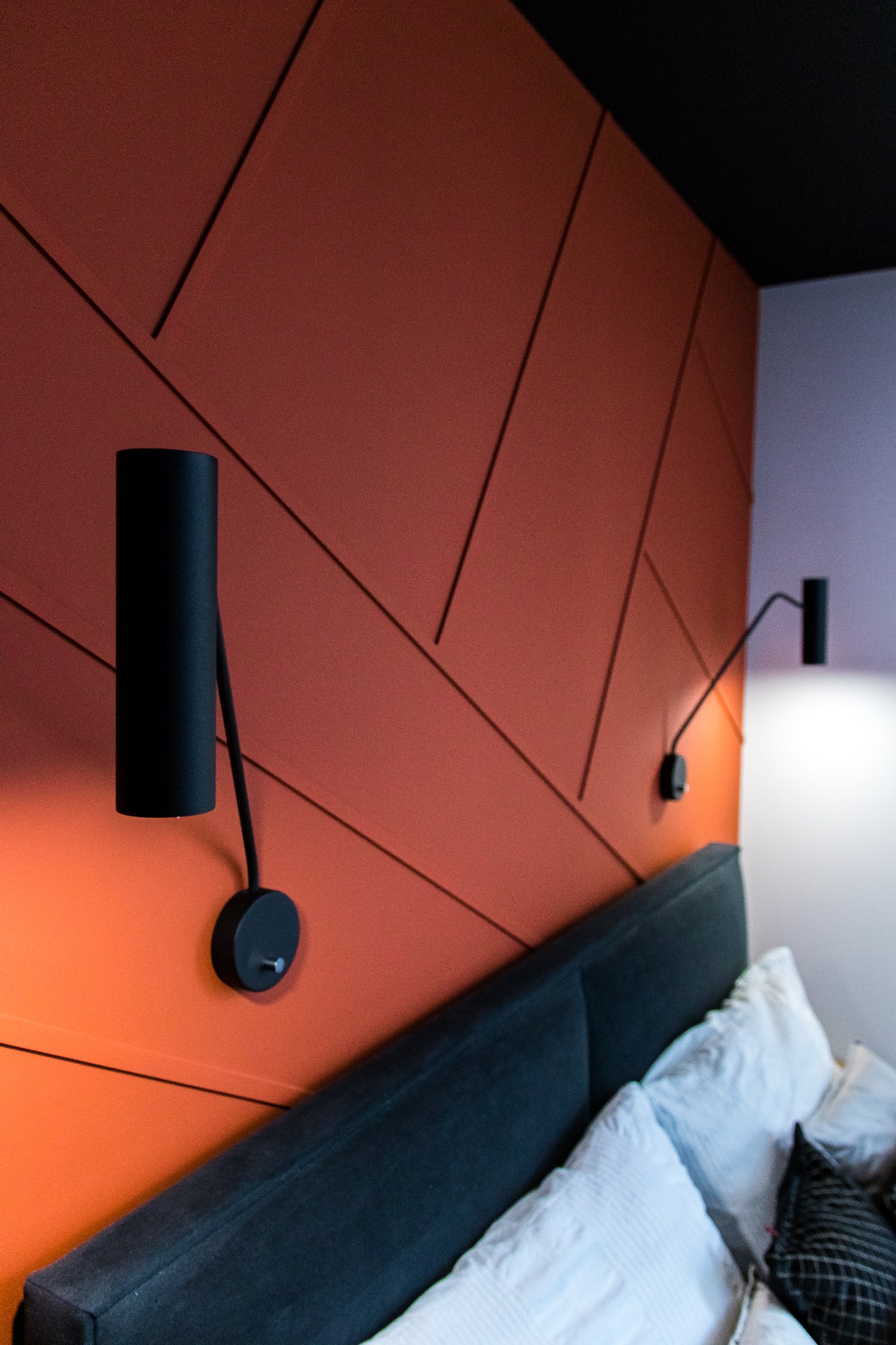 Pomarańczowa ściana w sypialni z geometrycznym wzorem z listew, czarne lampki nocne przy łóżku z szarym wezgłowiem i białymi poduszkami.