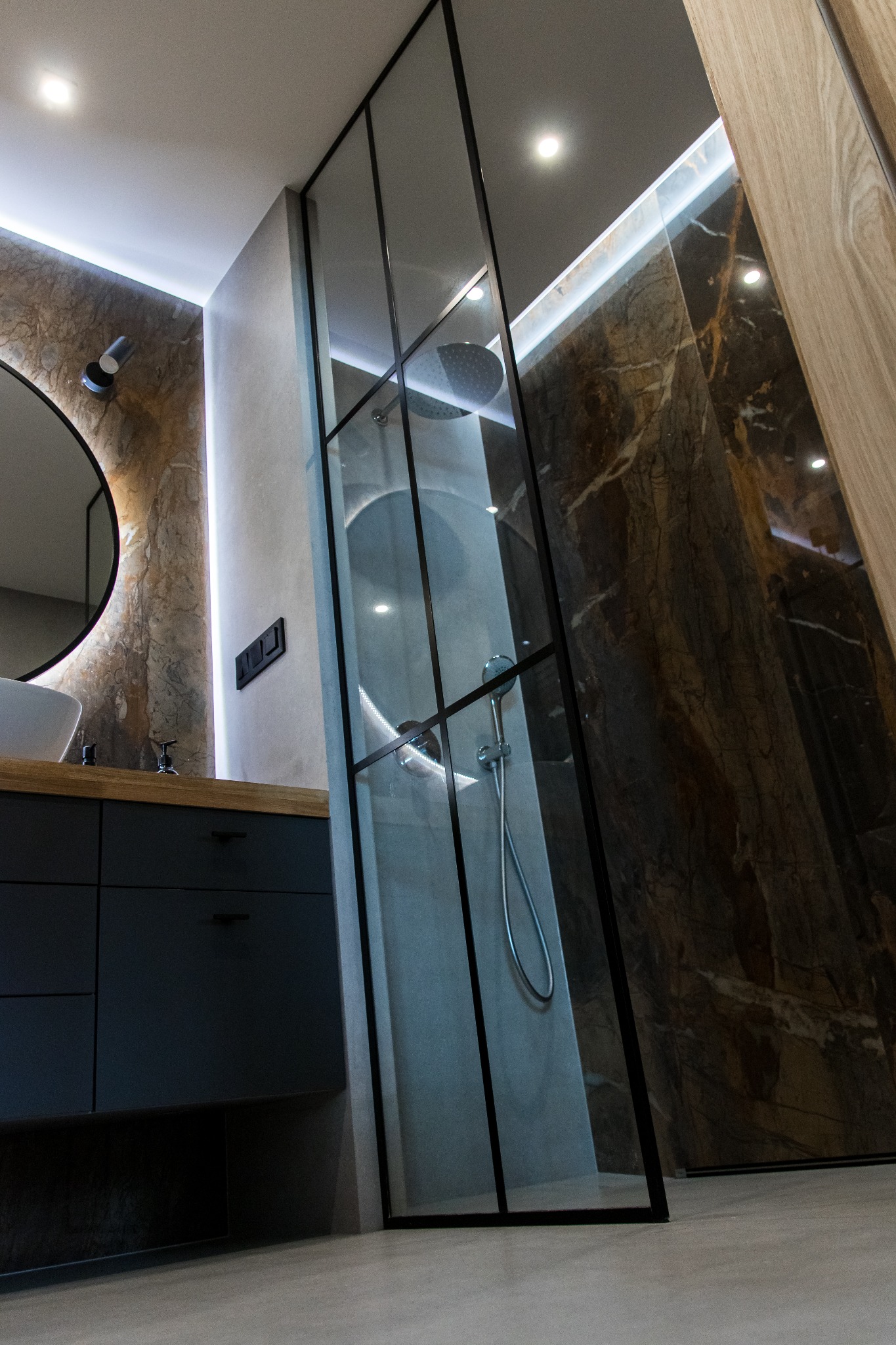 Nowoczesna łazienka z kabiną prysznicową typu walk-in, marmurowymi płytkami na ścianach, oświetleniem LED i umywalką nablatową na ciemnej szafce.