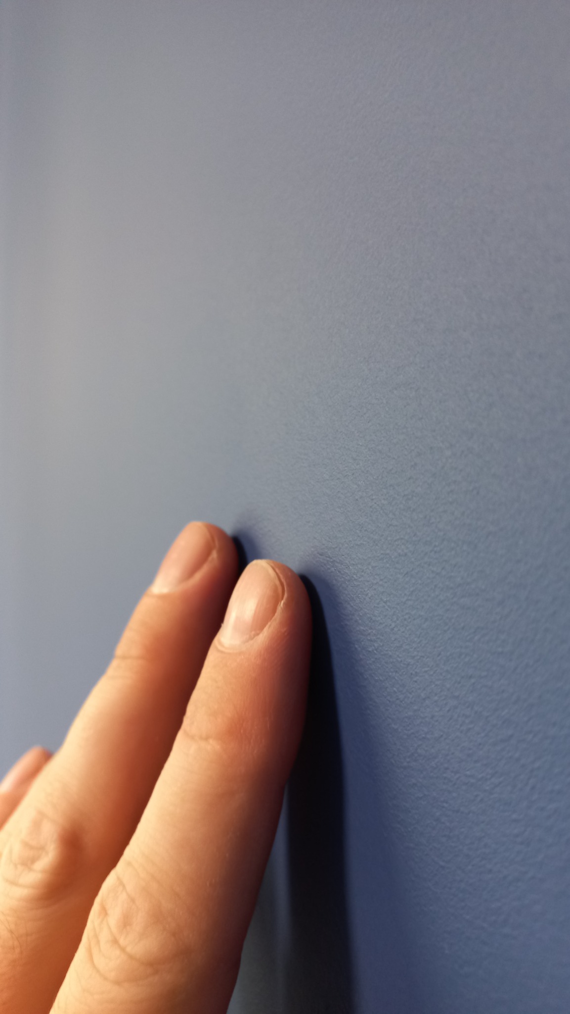 Dwa palce dotykają świeżo pomalowanej, gładkiej ściany w odcieniu niebieskiego, ukazując jej fakturę.