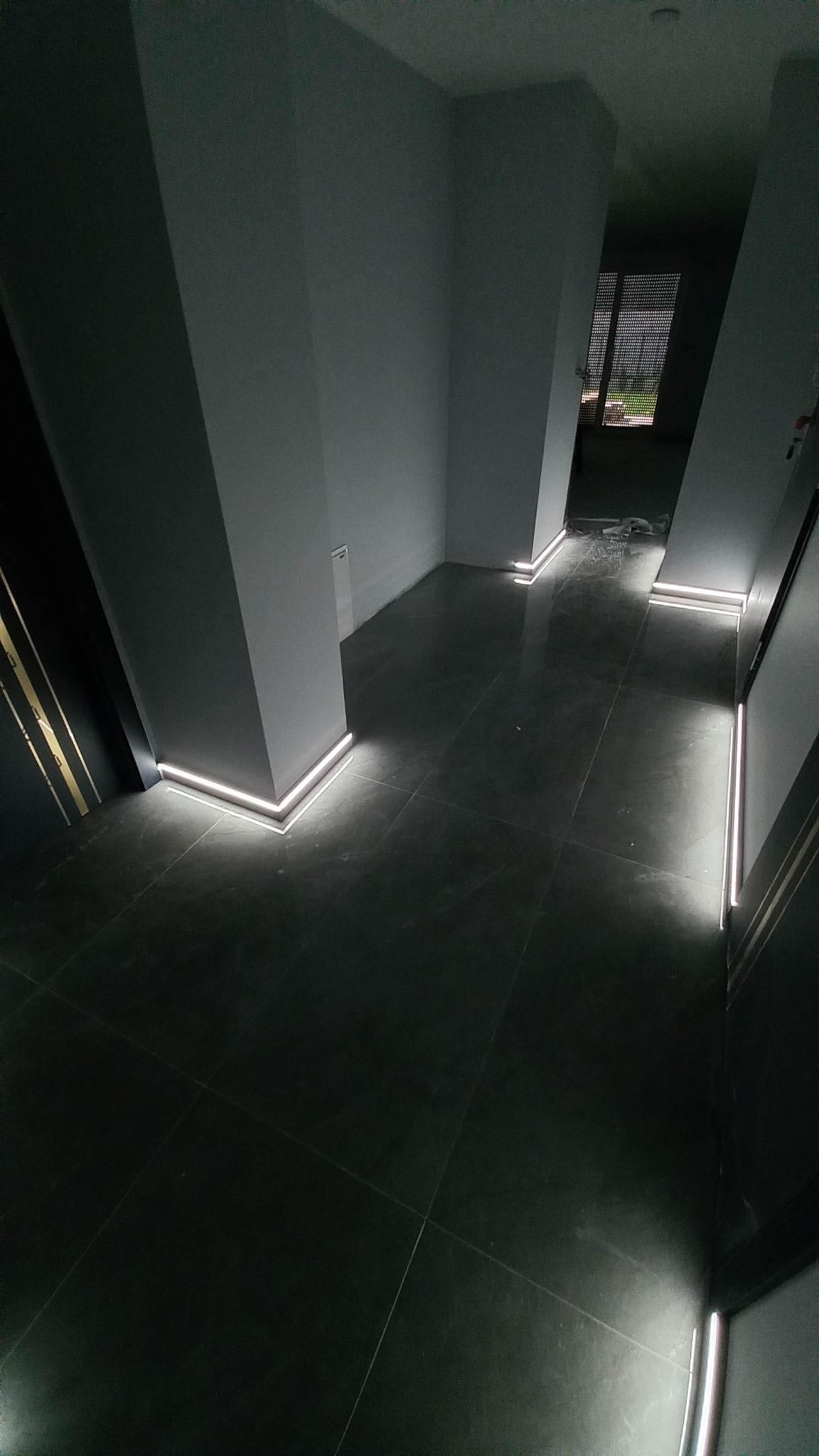 Korytarz z szarą, kwadratową podłogą i oświetleniem LED przy listwach przypodłogowych, widoczne słupy konstrukcyjne i fragment okna w oddali.