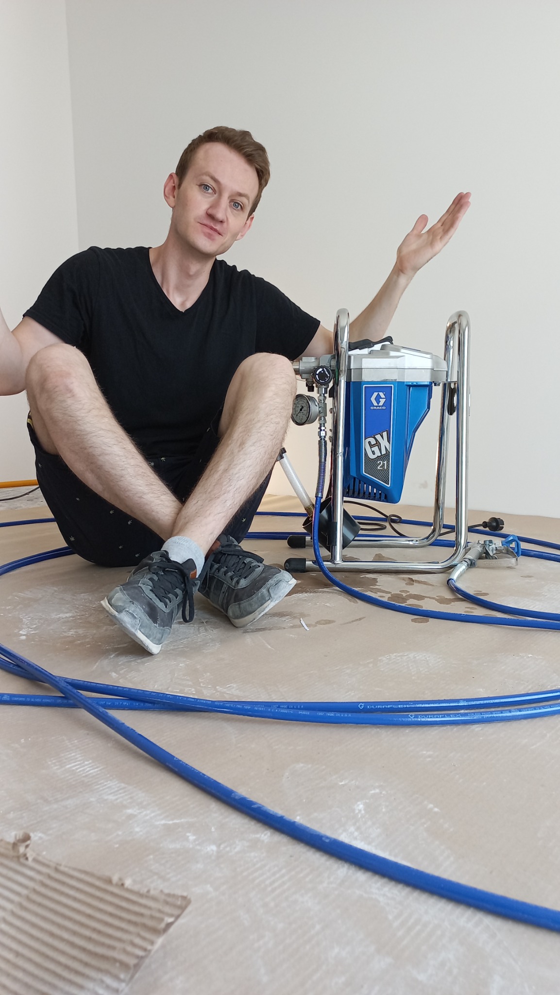 Mężczyzna w czarnej koszulce siedzi na podłodze obok profesjonalnego, niebieskiego agregatu malarskiego Graco GX 21, z rozłożonymi niebieskimi wężami, wskazując dłonią na urządzenie w pomieszczeniu...
