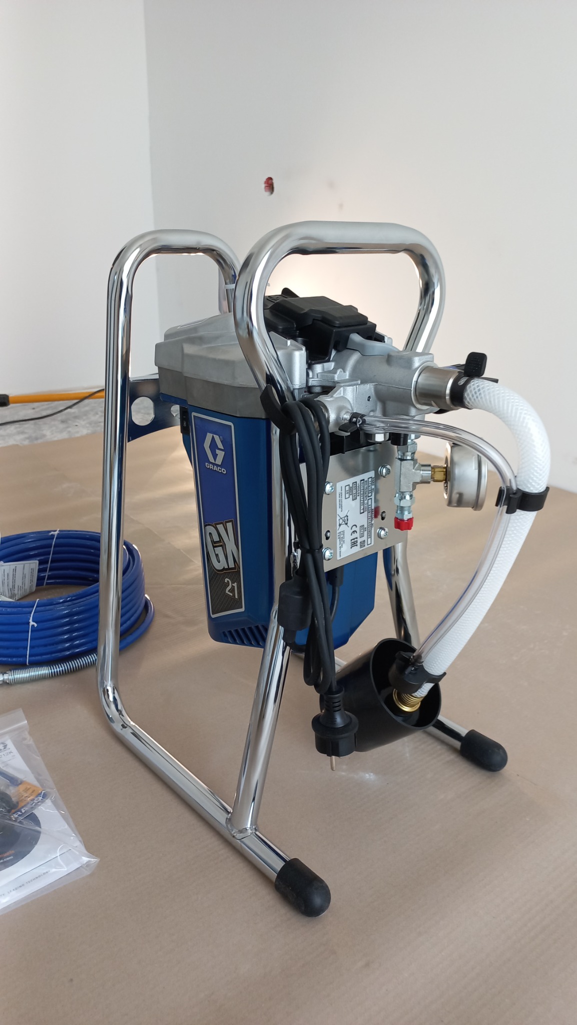 Profesjonalny agregat malarski Graco GN 21 na metalowym stelażu, z niebieską obudową i białymi wężami, stojący na zabezpieczonej podłodze w jasnym wnętrzu, gotowy do użycia.