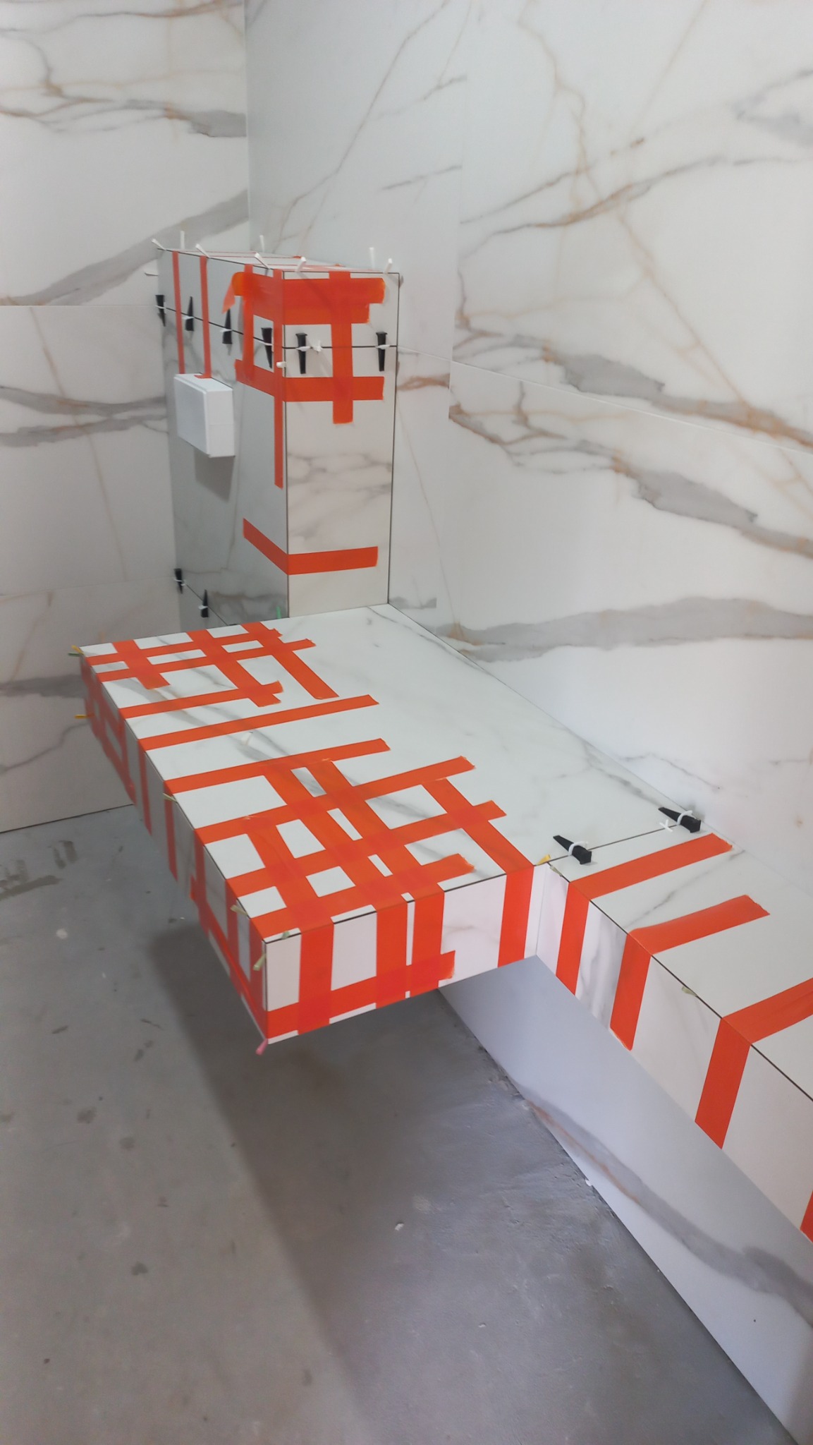 Montaż płytek imitujących marmur na zabudowie umywalki i ścianie, zabezpieczonych pomarańczową taśmą i poziomowane plastikowymi krzyżykami.