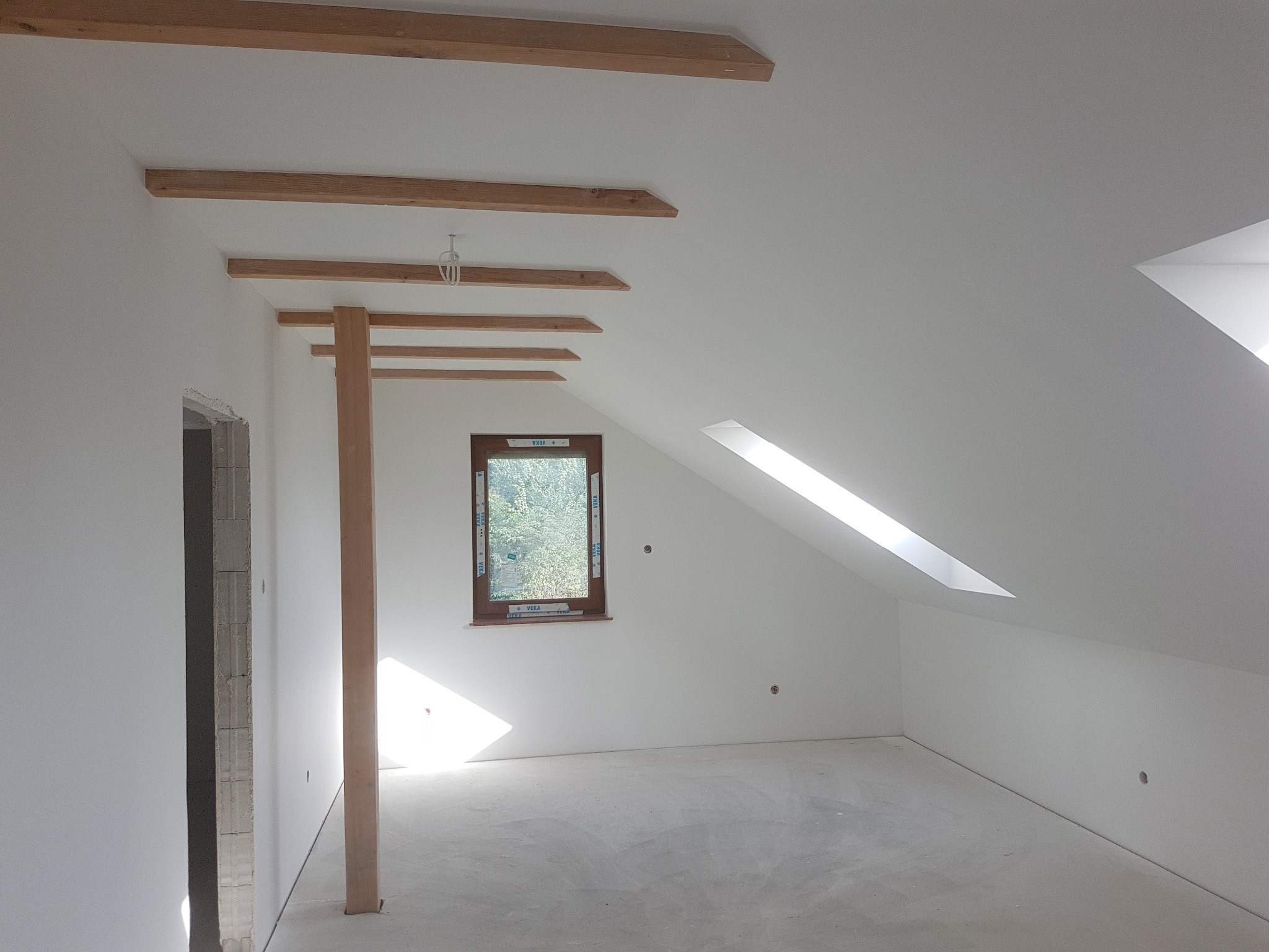 Poddasze w trakcie prac wykończeniowych: ściany pomalowane na biało, widoczne drewniane belki konstrukcyjne, okno dachowe i otwór drzwiowy w stanie surowym.