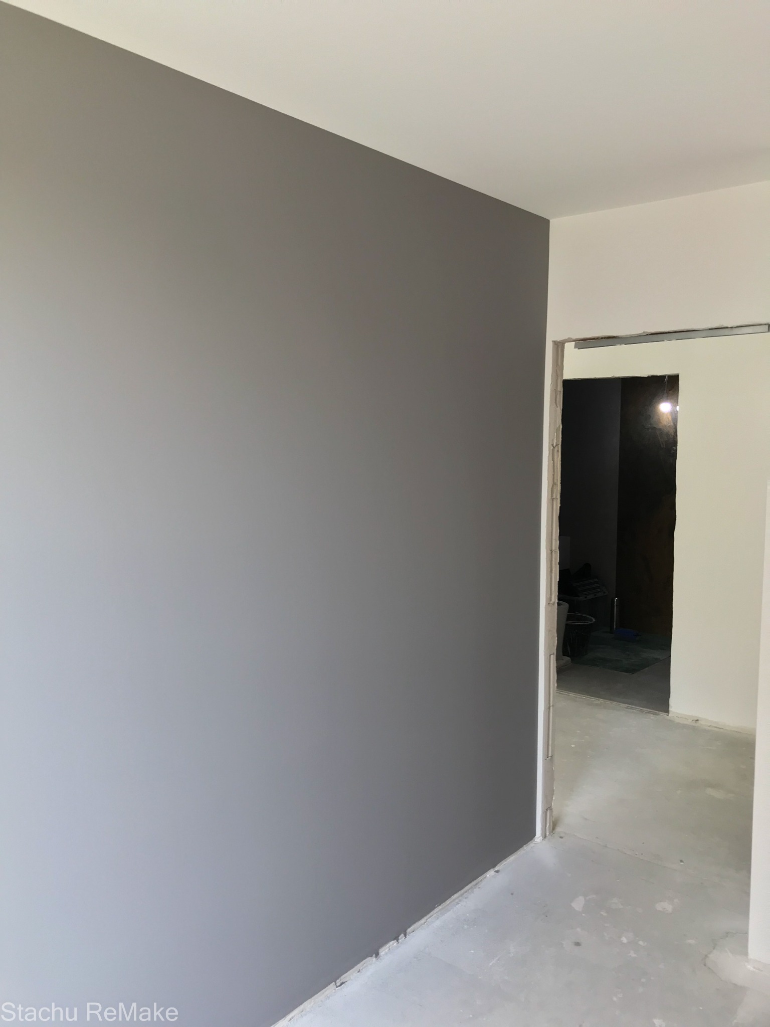 Świeżo pomalowana szara ściana w pokoju, widoczny surowy beton na podłodze i fragment otworu drzwiowego prowadzącego do ciemniejszego pomieszczenia z widocznymi narzędziami.