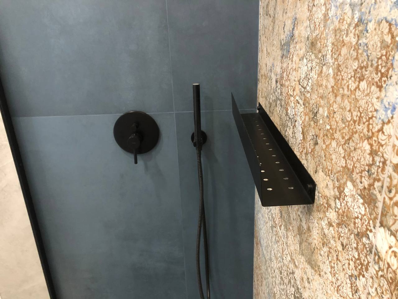 Nowoczesna łazienka z szarymi płytkami i czarną armaturą: bateria prysznicowa z deszczownicą i półka na kosmetyki na ścianie z dekoracyjnym wzorem.