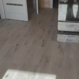 Ułożona podłoga z paneli imitujących drewno w jasnym odcieniu, widoczny fragment kominka wykończonego kamieniem dekoracyjnym i fragment ściany przygotowanej do malowania.