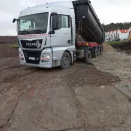 Srebrna ciężar&oacute;wka MAN z podniesioną skrzynią ładunkową wysypuje piasek na placu budowy, w tle budynki z czerwonymi dachami.