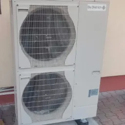 Zewnętrzna jednostka dwu-wentylatorowej pompy ciepła De Dietrich na betonowej podstawie, z widocznym logo producenta i fragmentem elewacji budynku w tle.