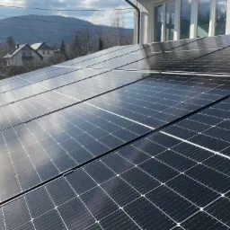 Część instalacji paneli słonecznych na dachu domu jednorodzinnego, widoczna osoba w czarnym ubraniu na skraju dachu, w tle góry i zabudowa mieszkalna.