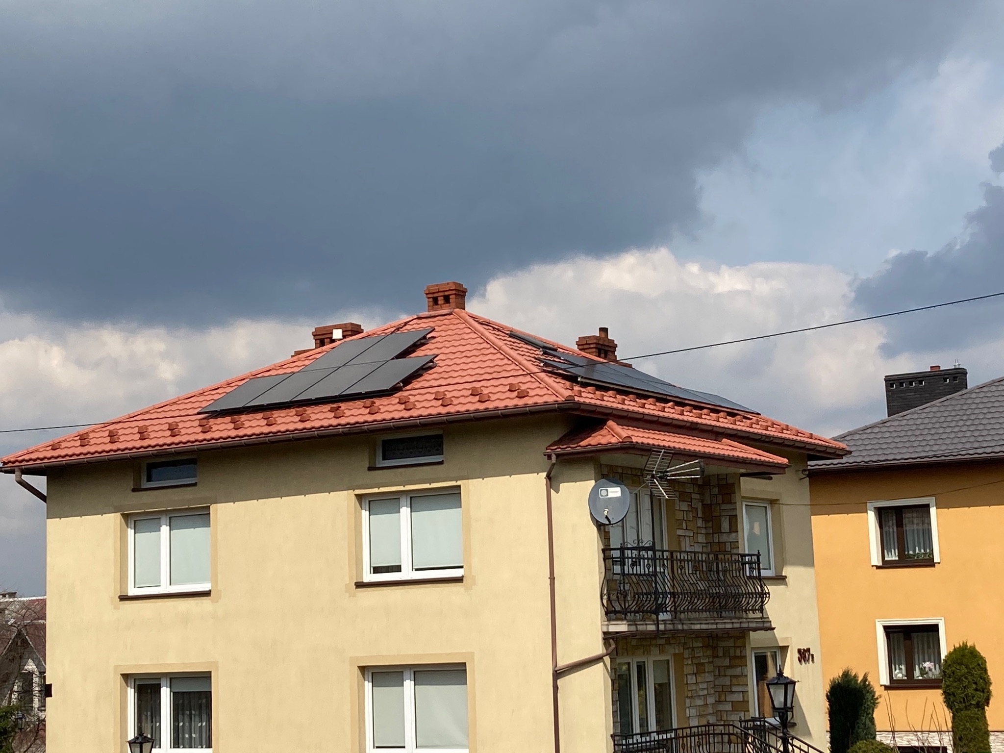 Dwupiętrowy dom z beżową elewacją i czerwoną dachówką, na której zamontowano panele słoneczne; widoczne czarne panele na tle burzowego nieba.