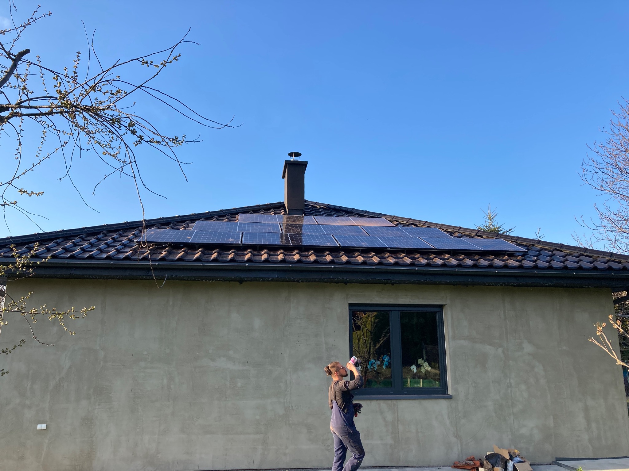 Nowoczesny dom jednorodzinny z panelami słonecznymi na dachu i pracownikiem w kombinezonie pijącym napój w pobliżu okna.