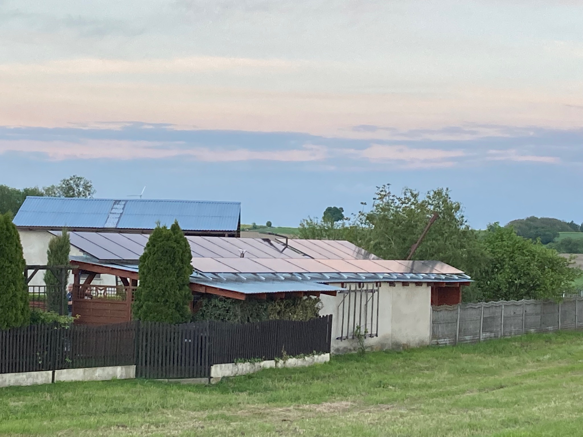 Dwa budynki z panelami słonecznymi na dachach, otoczone zielenią i ogrodzeniem. W tle widoczny krajobraz wiejski i zachmurzone niebo.