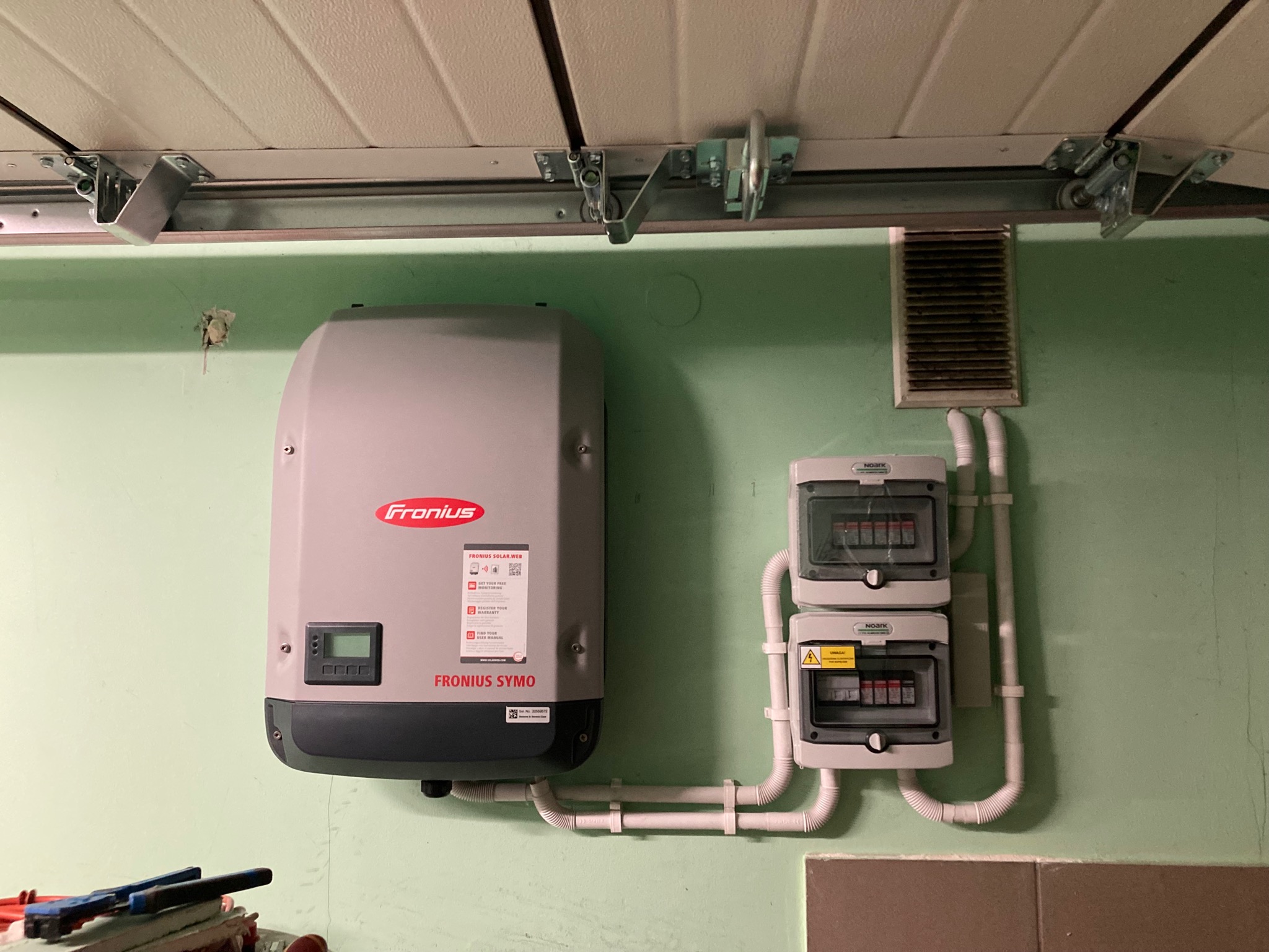Szary falownik Fronius Symo zamontowany na zielonej ścianie garażu, z widocznymi przewodami i skrzynkami elektrycznymi, oraz fragmentem narzędzi w lewym dolnym rogu.