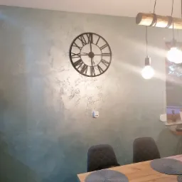 Ściana w salonie pomalowana farbą dekoracyjną w odcieniach szarości i zieleni, z dużym zegarem w stylu retro, nad drewnianym stołem z szarymi podkładkami i lampą wiszącą z drewnianym elementem.