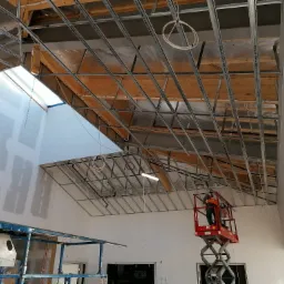 Montaż sufitu podwieszanego z płyt kartonowo-gipsowych, widoczne stalowe profile konstrukcyjne, oświetlenie tymczasowe i pracownik na podnośniku nożycowym, ściany częściowo pokryte gładzią gipsową.