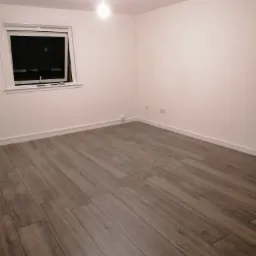 Pusty pokój z szarymi panelami podłogowymi ułożonymi wzdłuż długości pomieszczenia, białe ściany, okno z białymi ramami i listwa przypodłogowa w tym samym kolorze, centralnie umieszczona lampa...