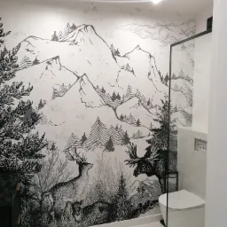 Wzorzysta tapeta w łazience przedstawiająca górski krajobraz z jeleniami i łosiem, połączona z nowoczesną białą armaturą i szklaną kabiną prysznicową.