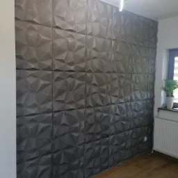 Ściana wykończona panelami 3D o geometrycznym wzorze w odcieniach szarości, oświetlona nowoczesną lampą sufitową, z widocznymi drewnianymi panelami podłogowymi.