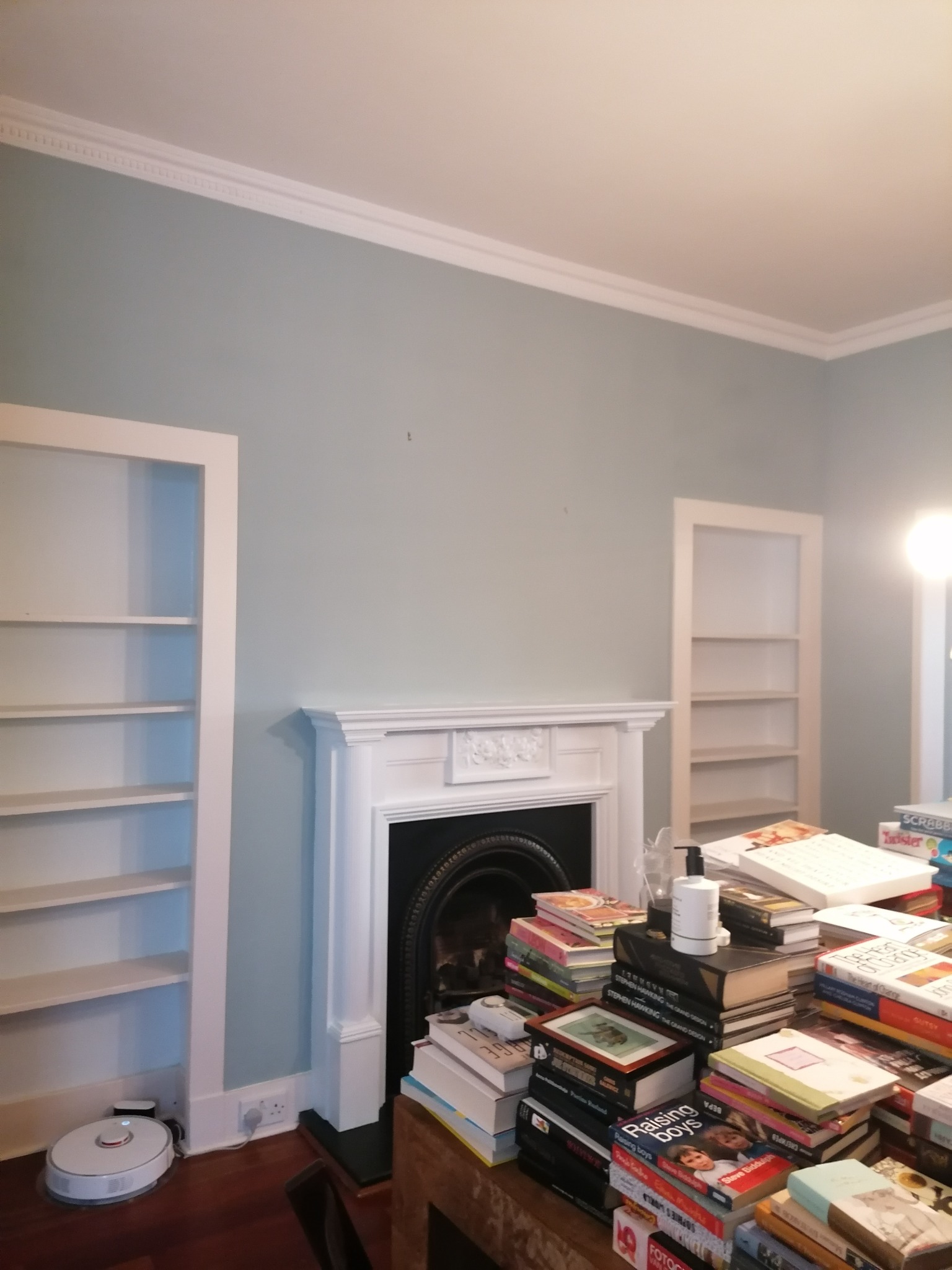 Pokój z pomalowanymi na jasno szaro ścianami, białym sufitem z dekoracyjną listwą, białym kominkiem i wbudowanymi półkami po bokach kominka, zagracony stertami książek na stole. W rogu widoczny...