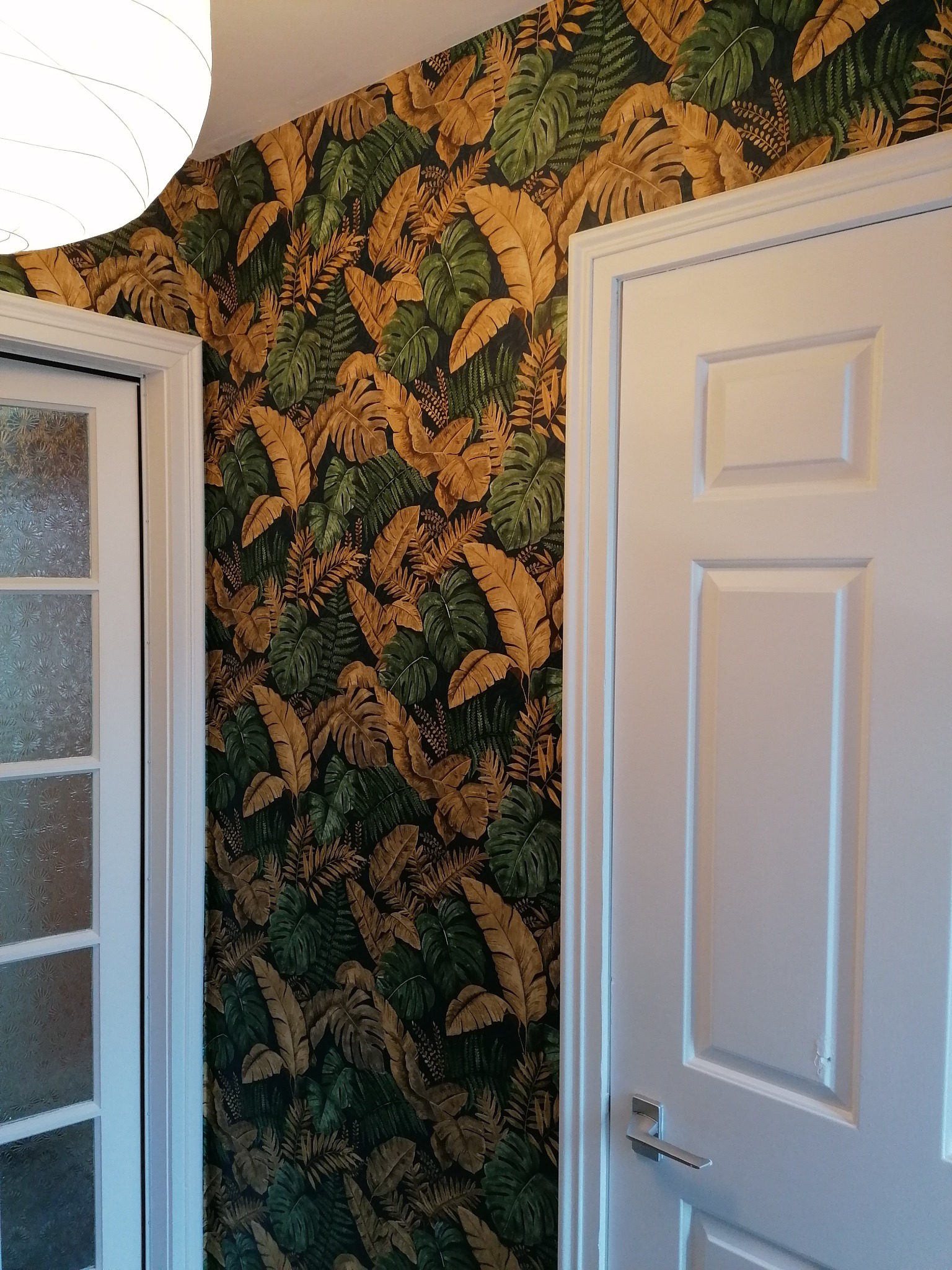 Ściana z tapetą w tropikalne liście, sąsiadująca z białą futryną drzwi i fragmentem lampy sufitowej.