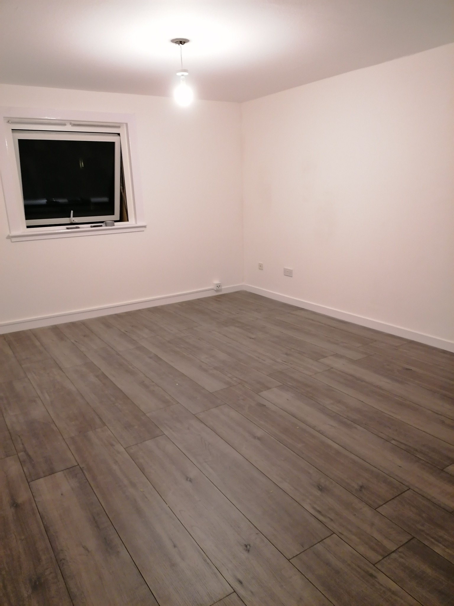 Pusty pokój z szarymi panelami podłogowymi ułożonymi wzdłuż długości pomieszczenia, białe ściany, okno z białymi ramami i listwa przypodłogowa w tym samym kolorze, centralnie umieszczona lampa...