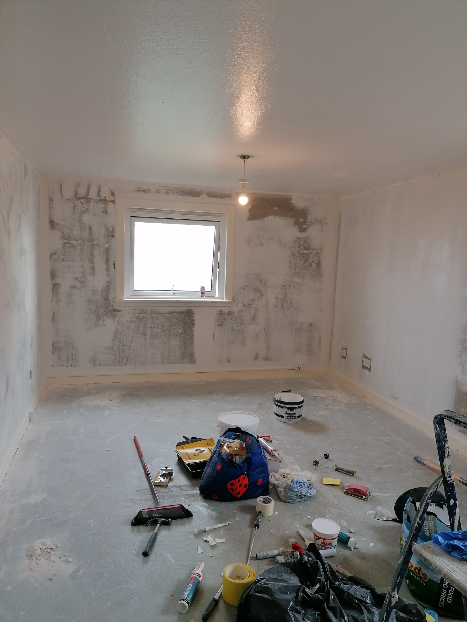 Pokój w trakcie przygotowań do malowania: ściany częściowo zagruntowane, widoczne ślady po szpachlowaniu, na podłodze rozłożone narzędzia i akcesoria malarskie, w tym pędzle, wałki, szpachelki...