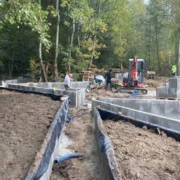 Wykop pod fundamenty budynku jednorodzinnego z widocznym szalunkiem, zabezpieczeniem folią i niebieską rurą drenażową; w tle pracownicy i minikoparka.