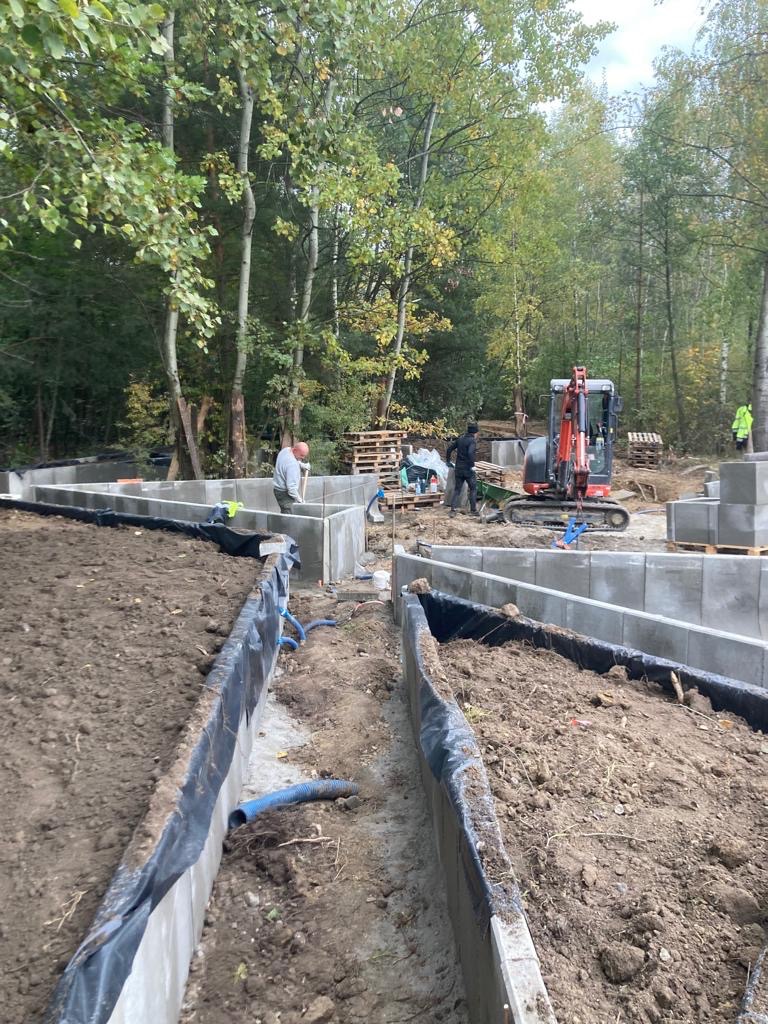 Wykop pod fundamenty budynku jednorodzinnego z widocznym szalunkiem, zabezpieczeniem folią i niebieską rurą drenażową; w tle pracownicy i minikoparka.