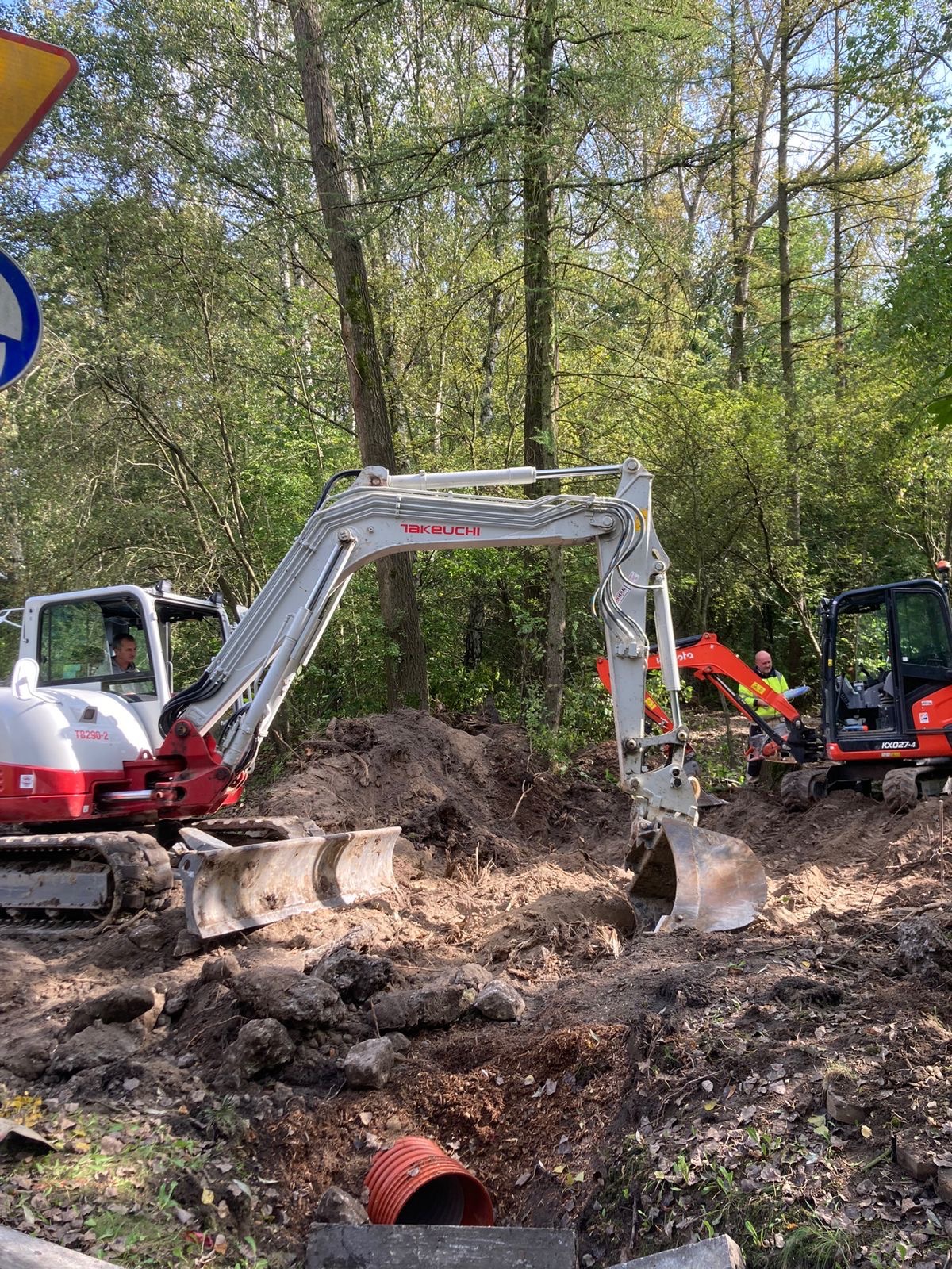 Dwa minikoparki Takeuchi i Kubota w trakcie prac ziemnych na tle drzew, widoczny fragment rury drenarskiej w wykopie.