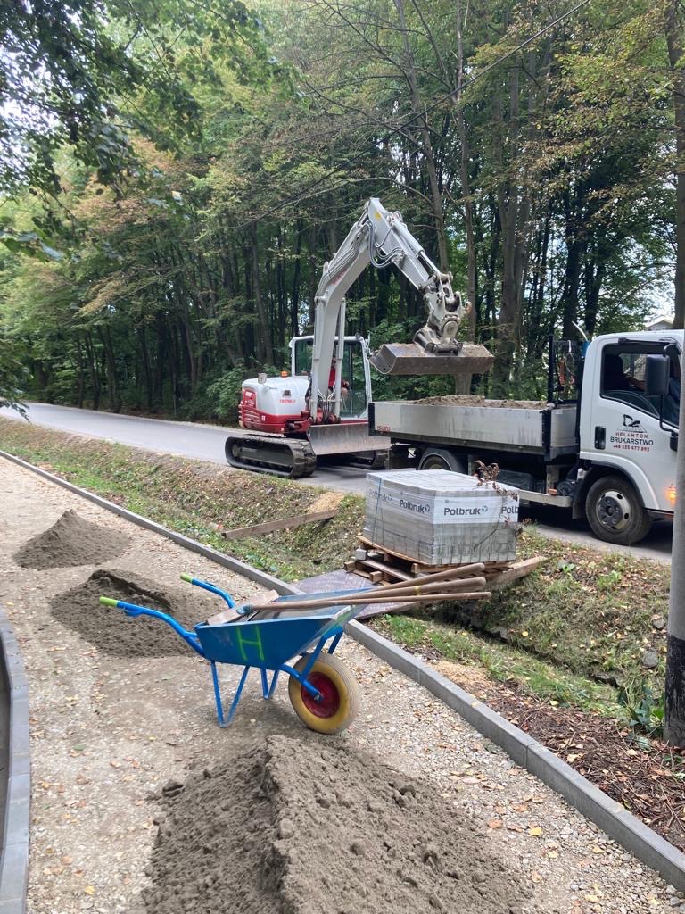 Przygotowanie terenu pod ścieżkę: mini koparka ładuje ziemię na ciężarówkę obok stosu kostki brukowej, taczki i narzędzi.