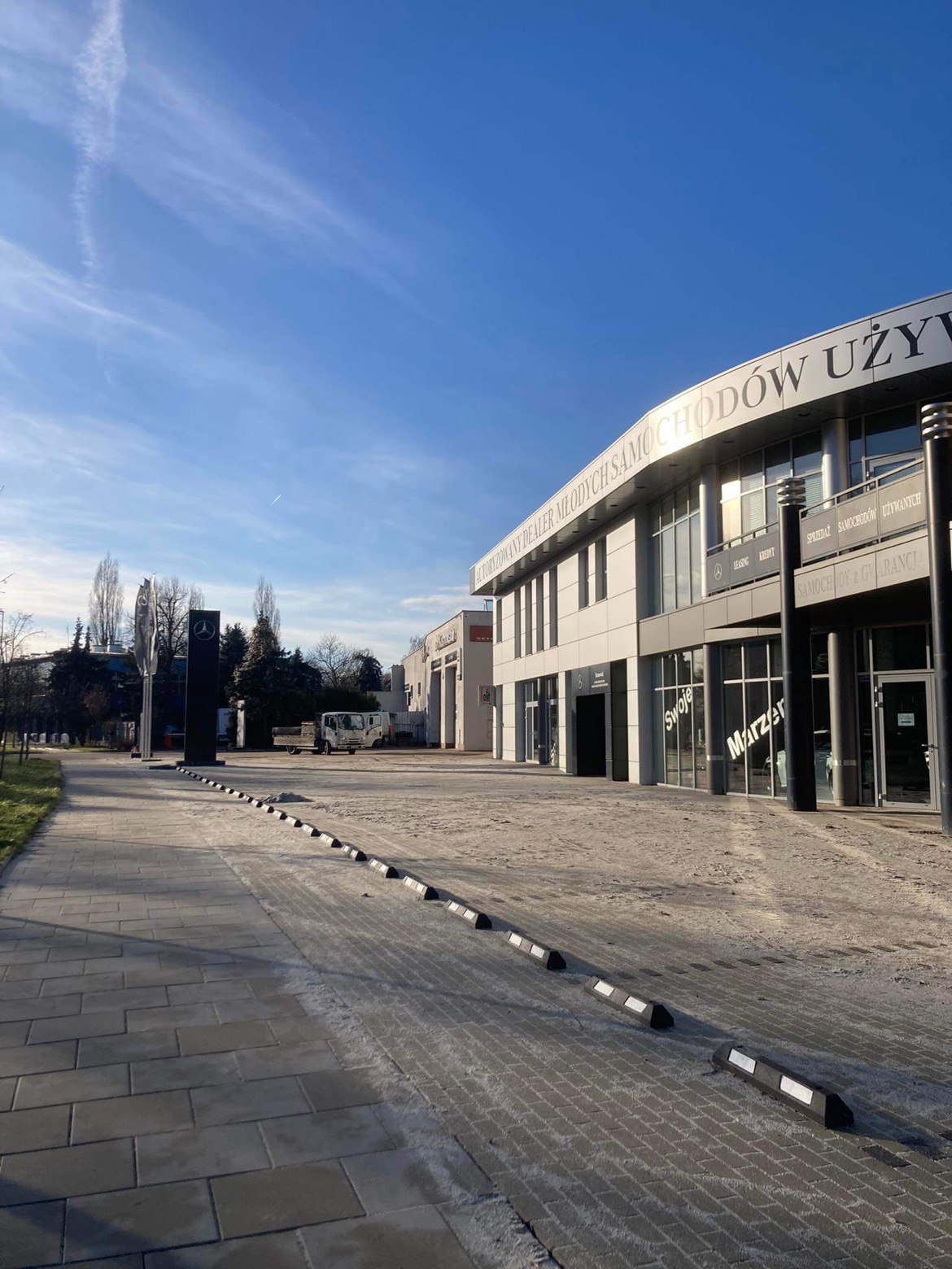 Szeroki kadr na plac przed salonem samochodowym z kostki brukowej z gumowymi ogranicznikami, widok na budynek z dużymi oknami i napisem 'AUTORYZOWANY DEALER MŁODYCH SAMOCHODÓW UŻYWANYCH'.