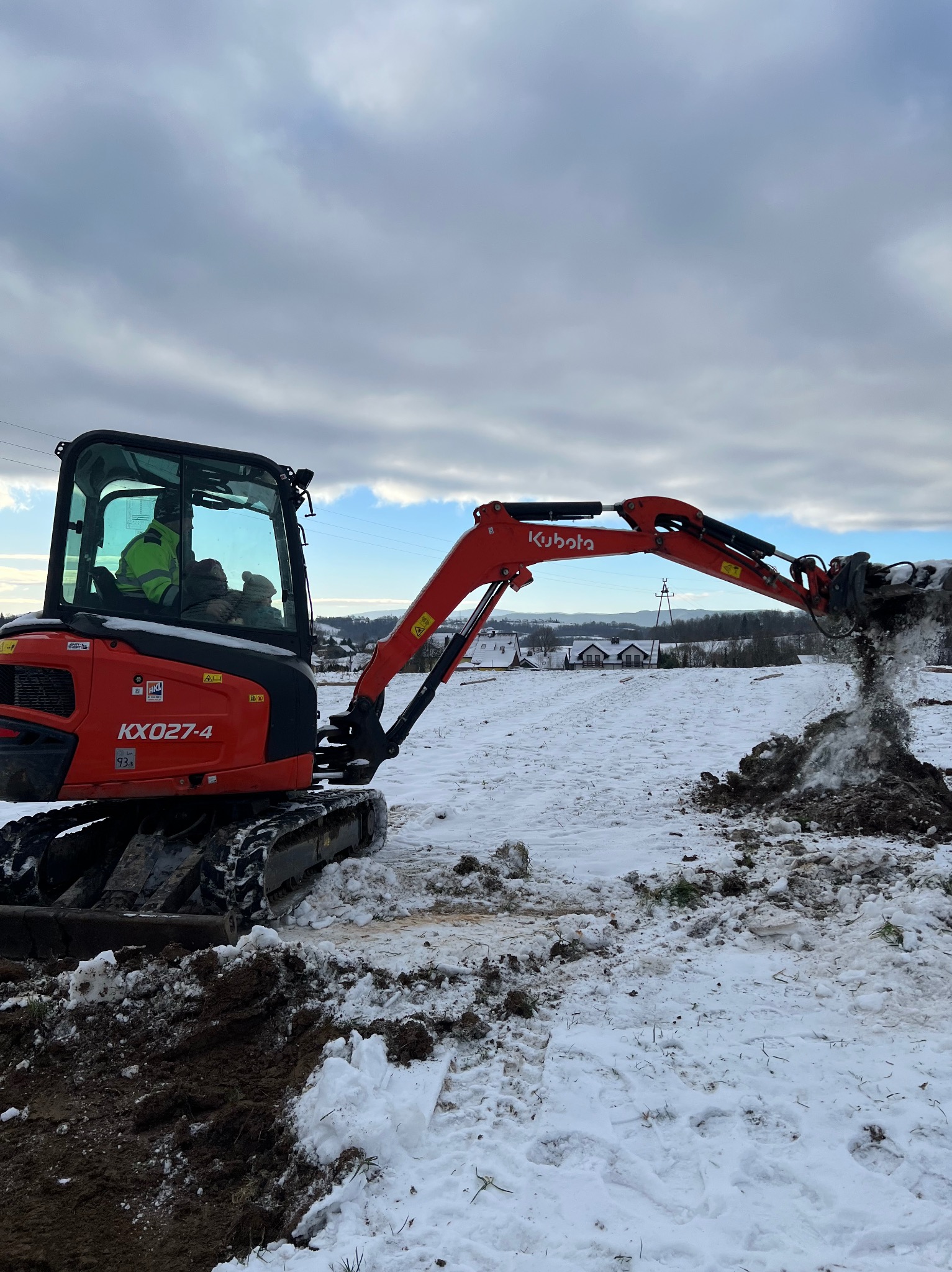 Czerwona mini koparka Kubota KX027-4 w trakcie pracy na zasypanym śniegiem terenie, wyrzucająca ziemię z wykopu, w tle zabudowania wiejskie i pochmurne niebo.